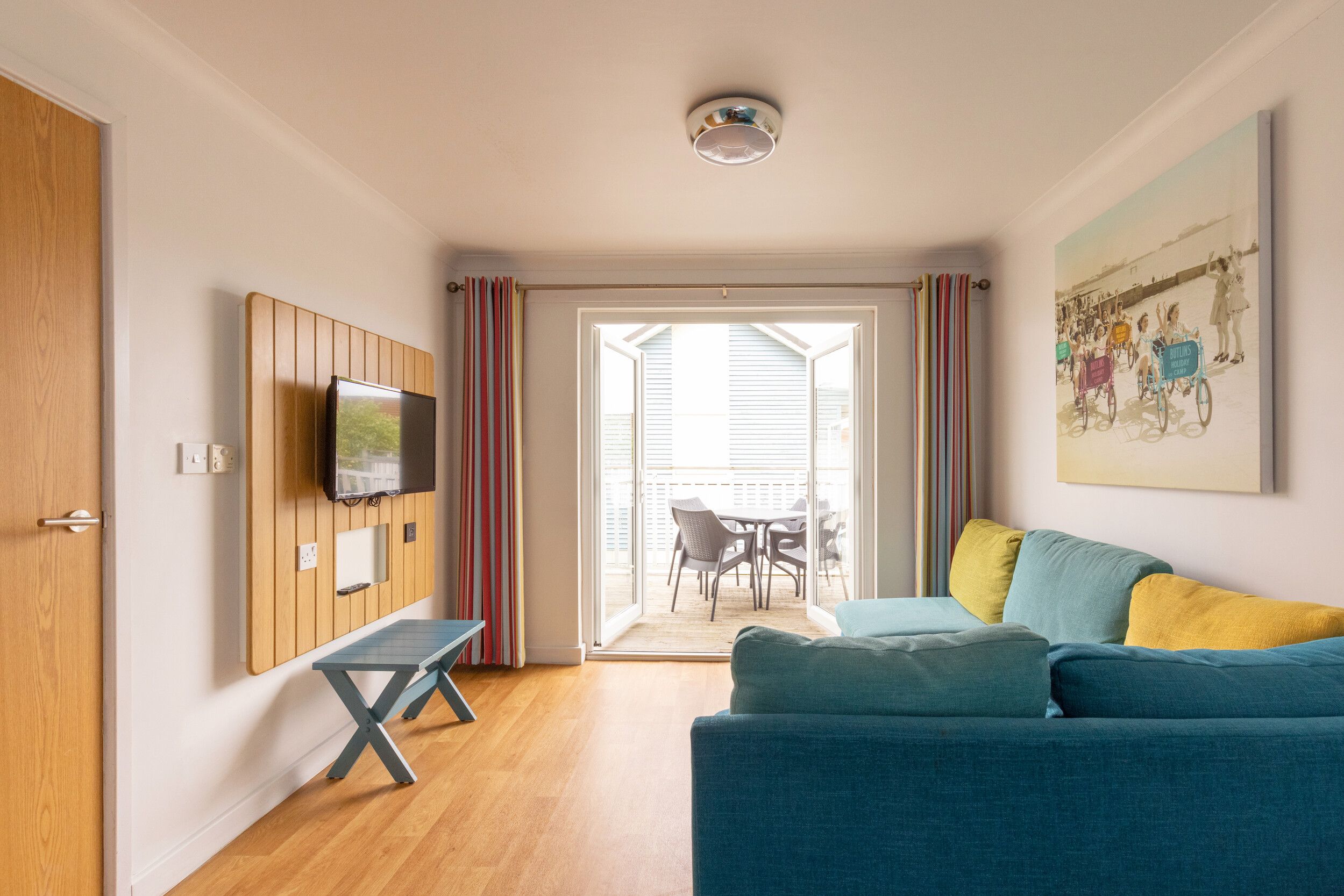 A living room area with a blue corner sofa in our Comfort Plus Apartments in Bognor Regis. There is a TV on the wall opposite with a small table underneath it. In the background you can see doors leading out into a terrace or balcony.