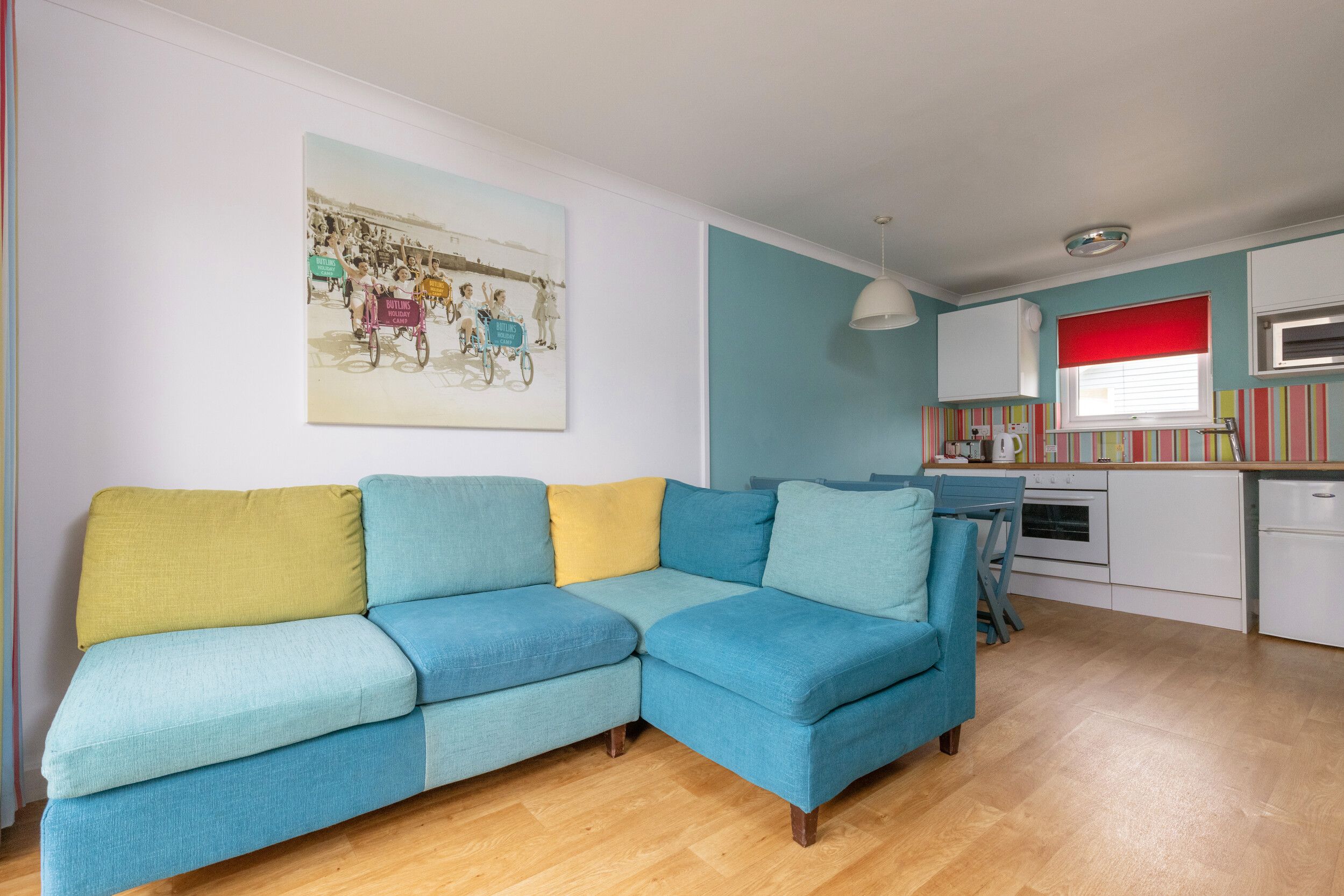 A living room with blue and yellow corner soft in our Comfort Plus Apartments at Bognor Regis. There is a small kitchen behind the sofa with an oven and microwave.