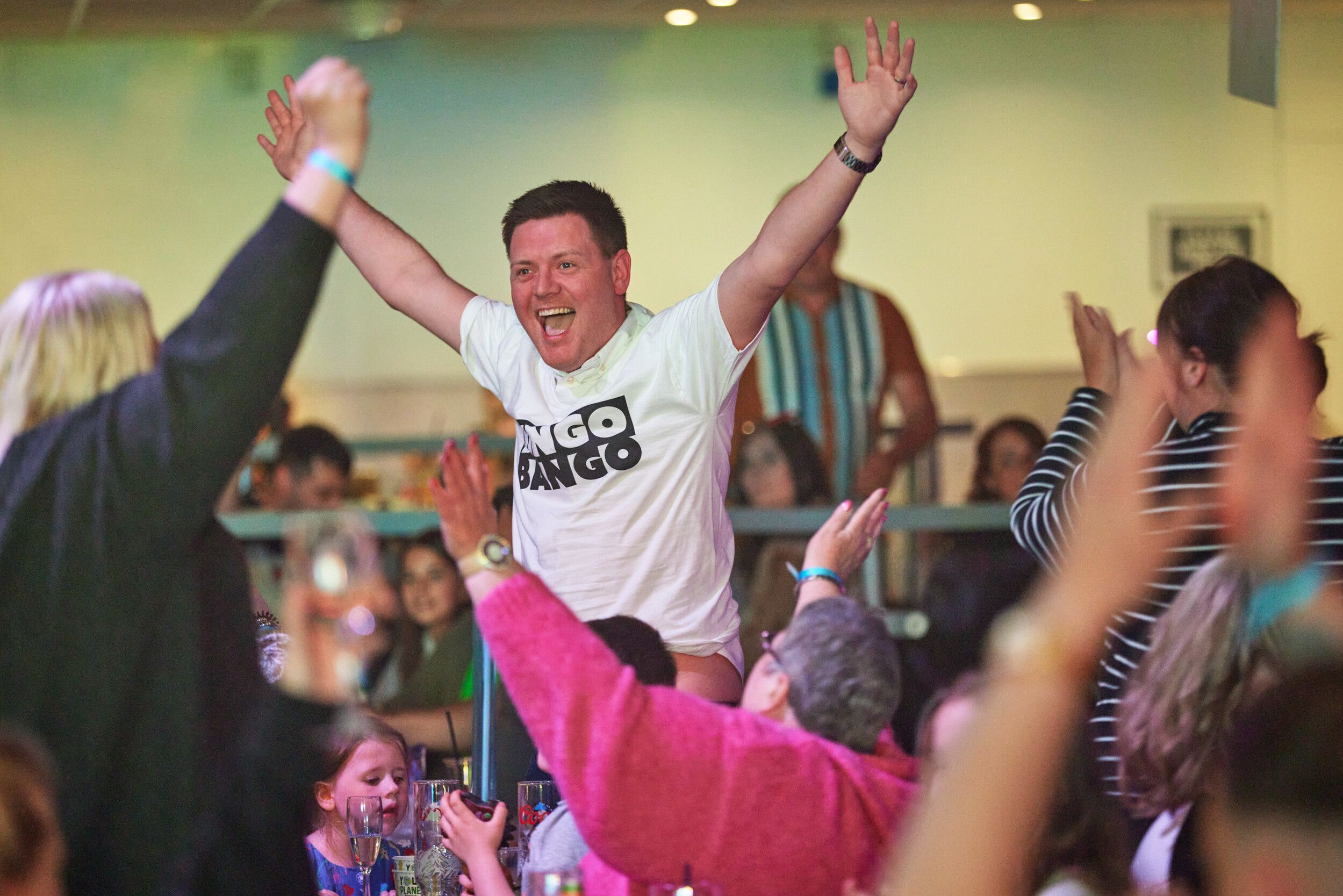 A person with a white "Bingo Bango" shirt, arms raised in celebration at an event.