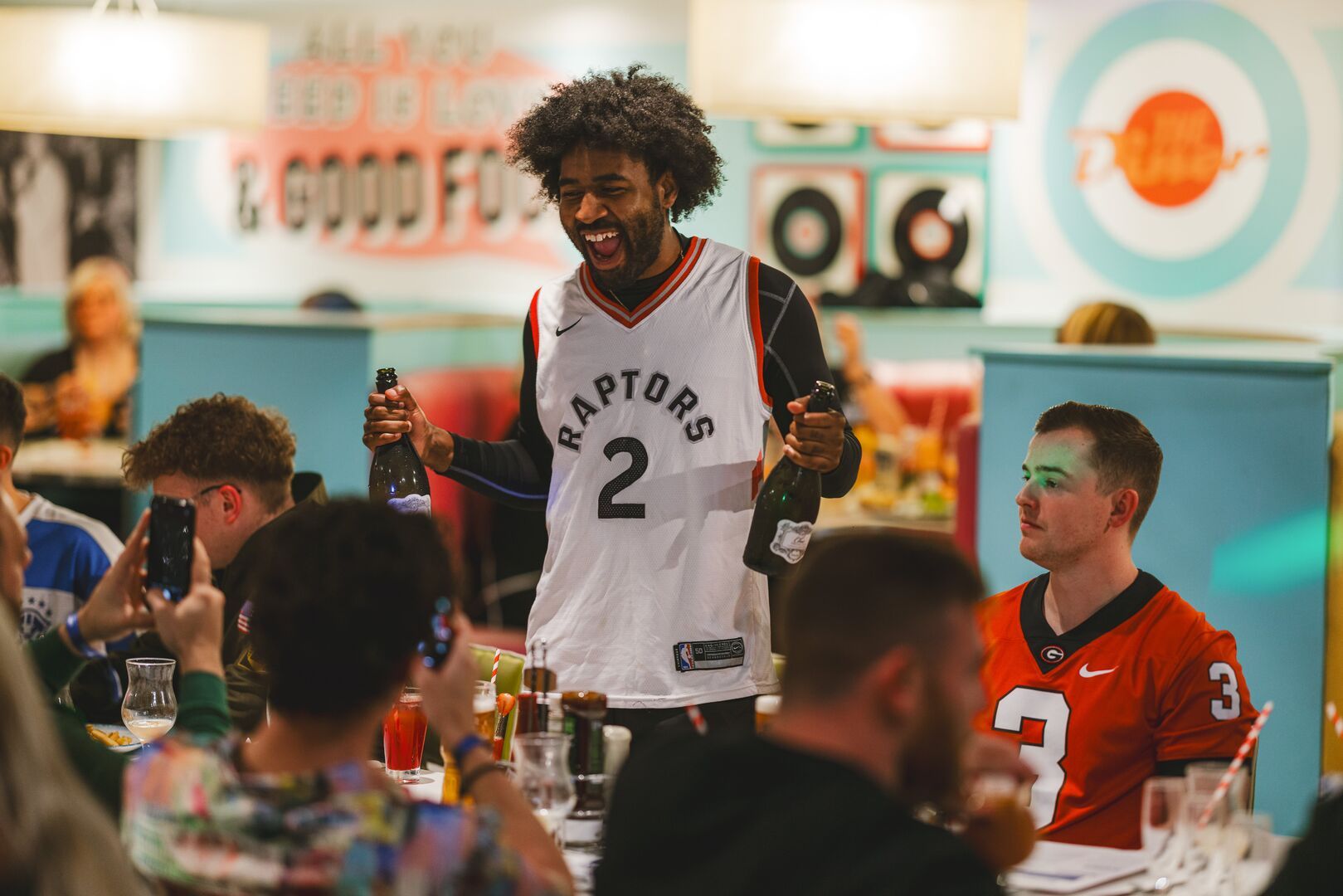 A group of friends enjoy a Bottomless Brunch session together during a Butlin's Big Weekender. One member of the group poses for a photo with a bottle of wine in his hand, as he looks at their cameras excitedly.