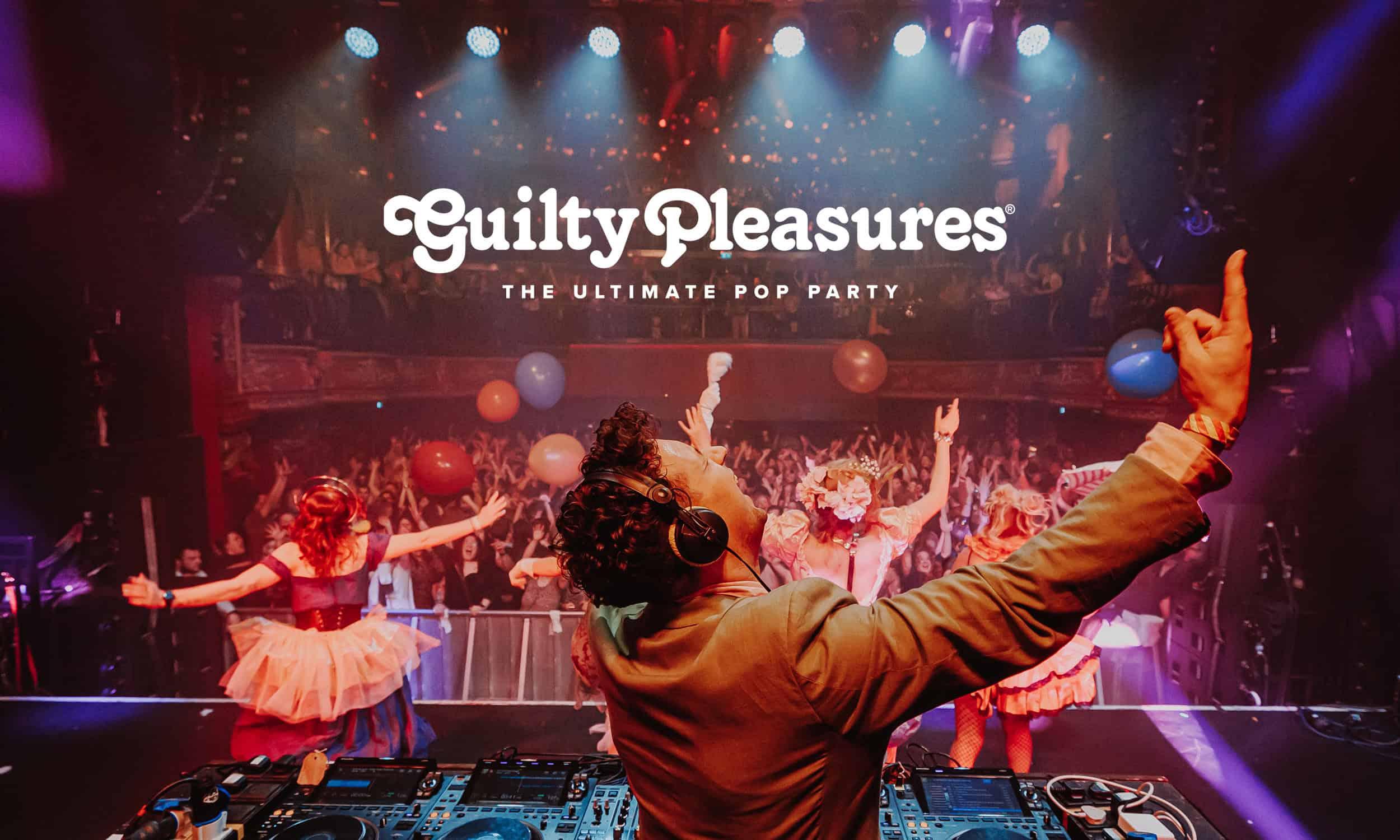 A DJ with arms raised celebrates in front of a packed, energetic crowd with colourful balloons and costumed dancers at a Guilty Pleasures club night, branded as "The Ultimate Pop Party"