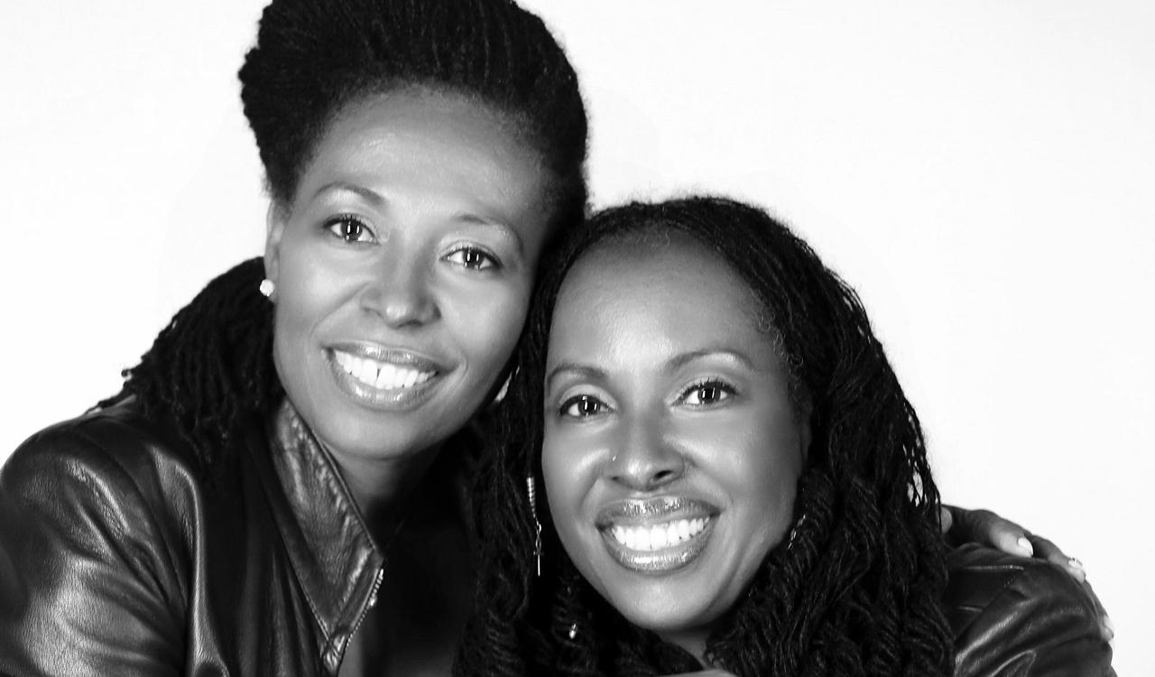 Carroll Thompson and Janet Kay studio portrait. Black and white close-up of two smiling women with their heads together and an arm around each other, both with long locs and wearing leather jackets, against a plain white background.