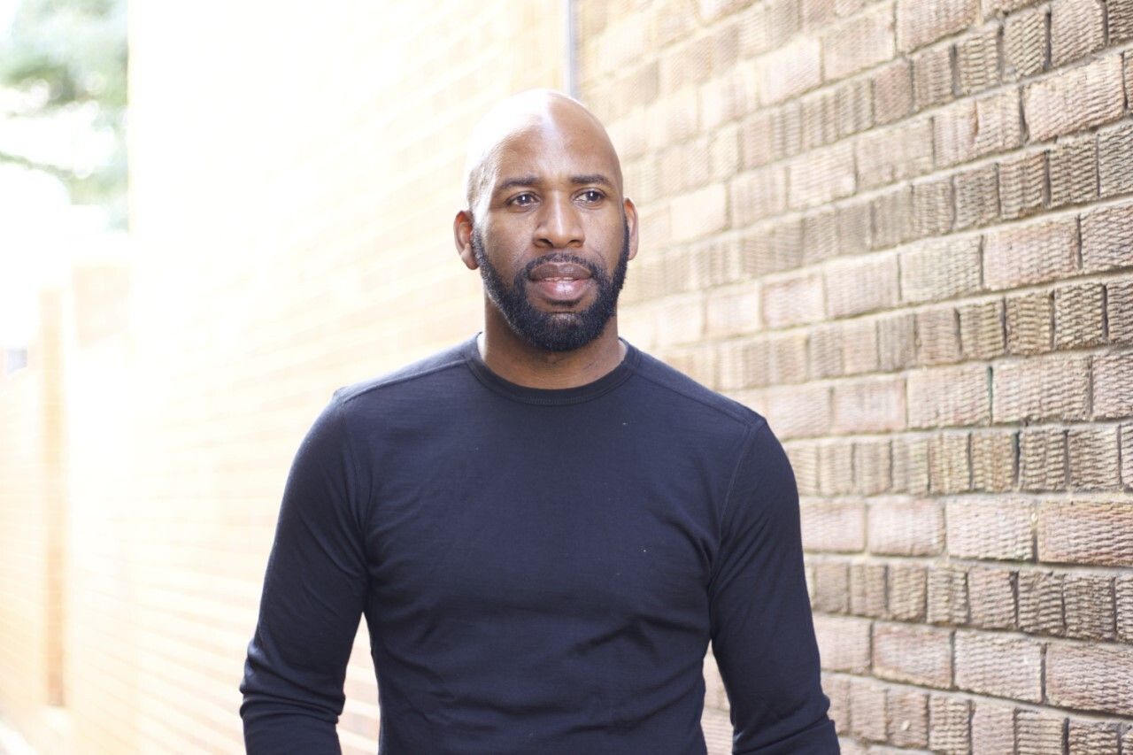 DJ Spoony standing in front of the camera beside a brown brick wall.