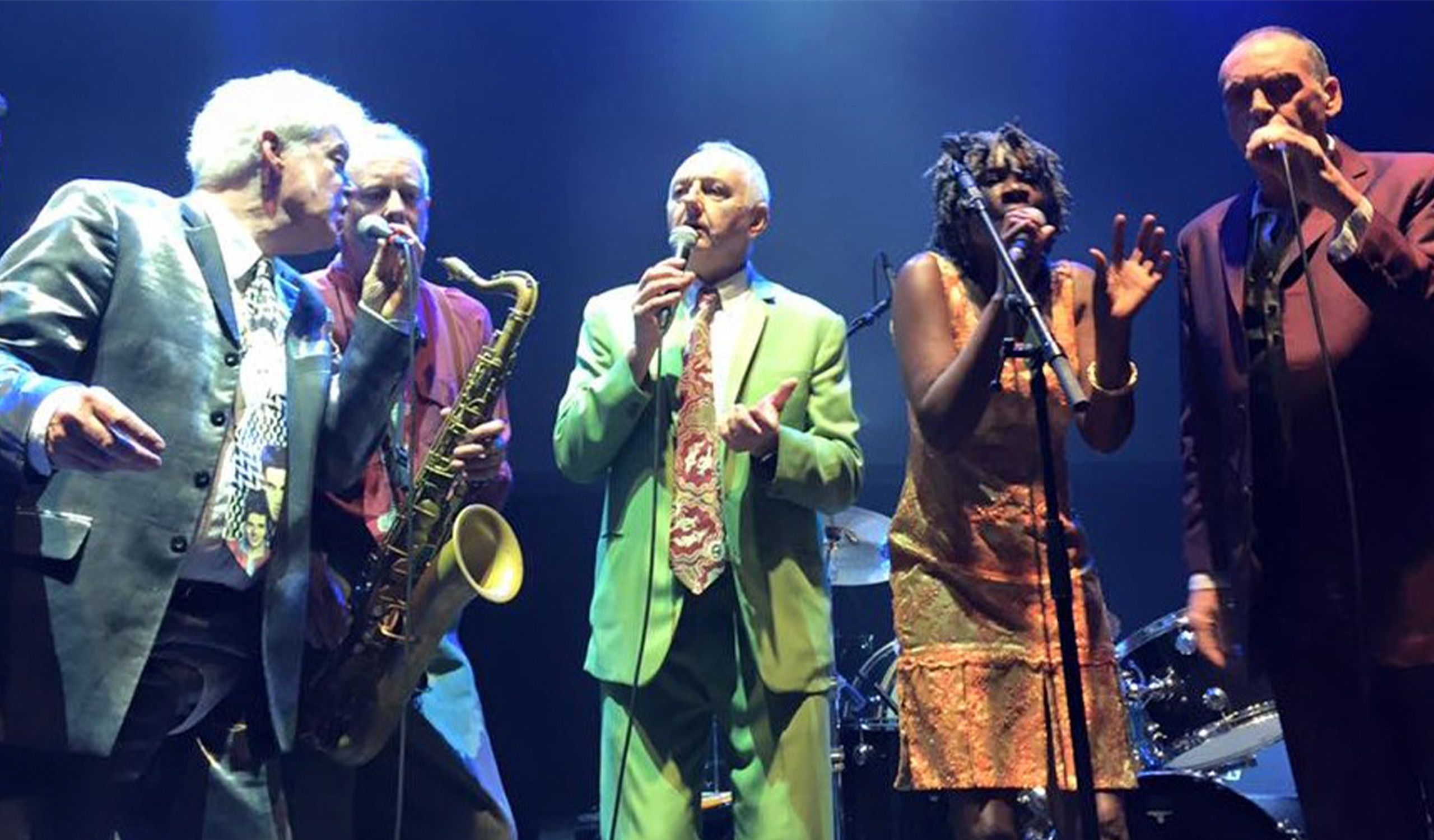 Members of the band Darts on stage, singing together with one playing saxophone.