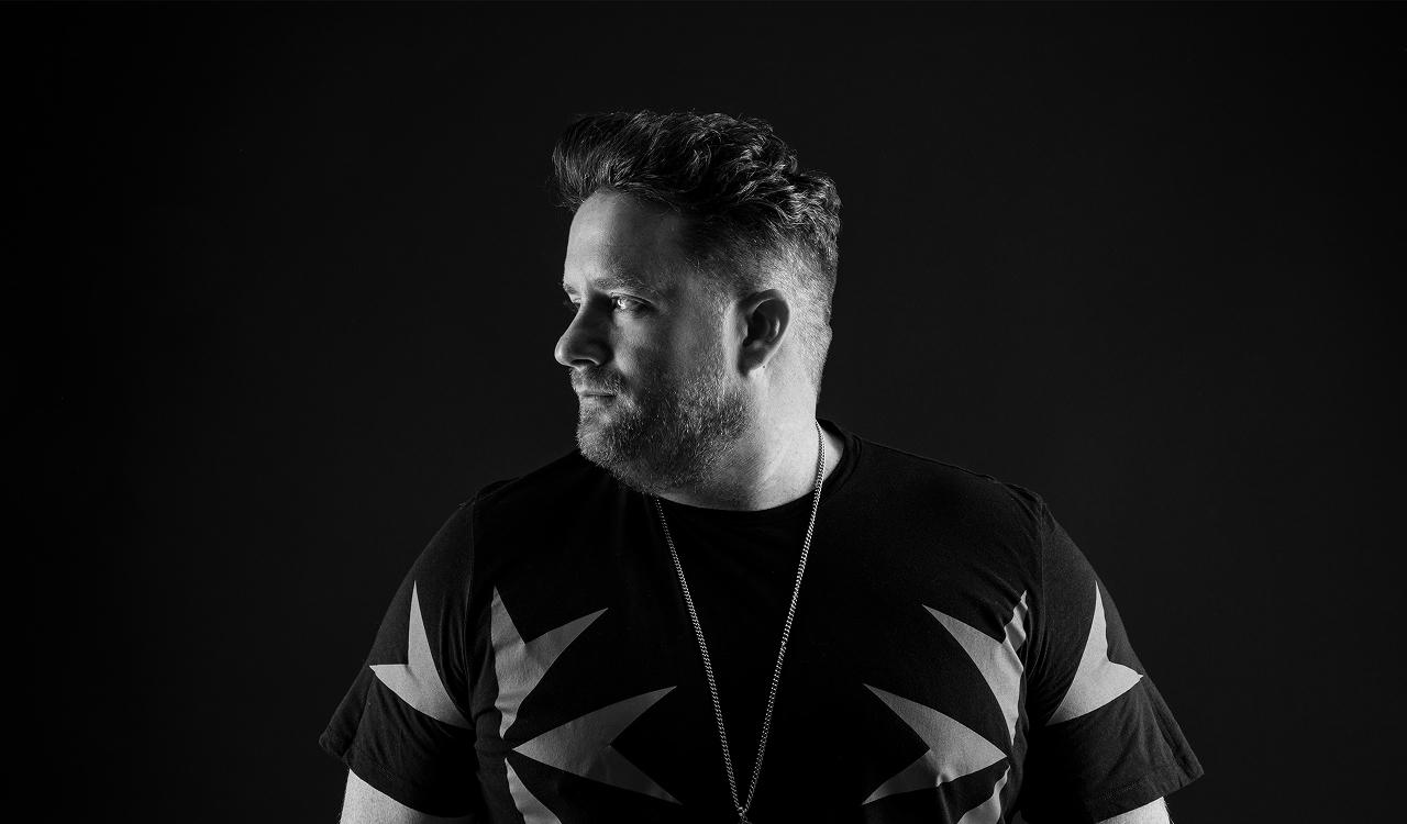 Eats Everything in a black and white portrait against a dark background, looking to the side, wearing a black t-shirt with a bold star pattern and a silver chain necklace.