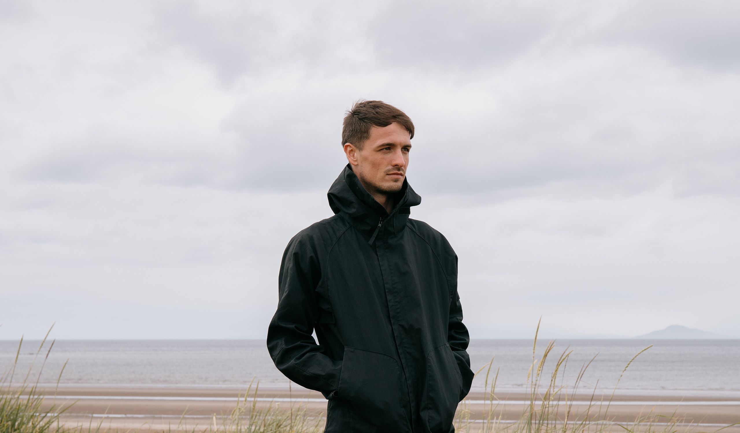 Artist Ewan McVicar standing in a dark jacket on the coast with the sea behind him.