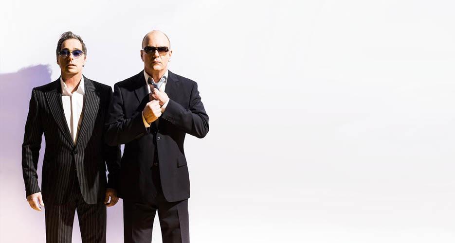 Go West, two men in dark suits and sunglasses standing side by side against a bright white background. The man on the left wears a pinstripe suit with an open-collared white shirt; the man on the right wears a plain dark suit and adjusts his tie with both hands. Open space to the right of the frame.