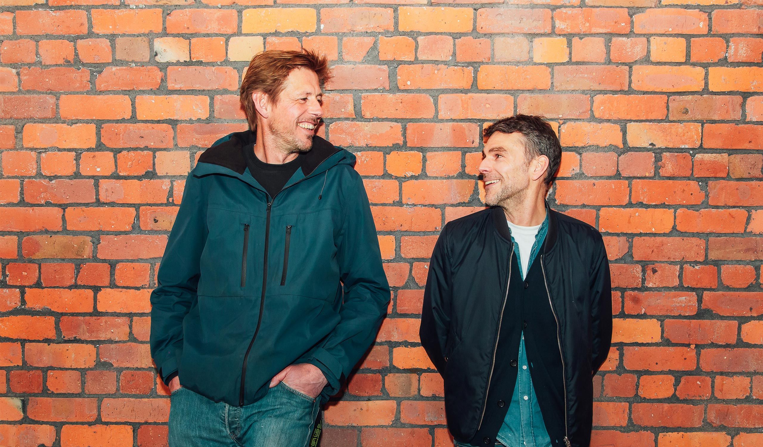 The wo members of Groove Armada laughing together against a wall.