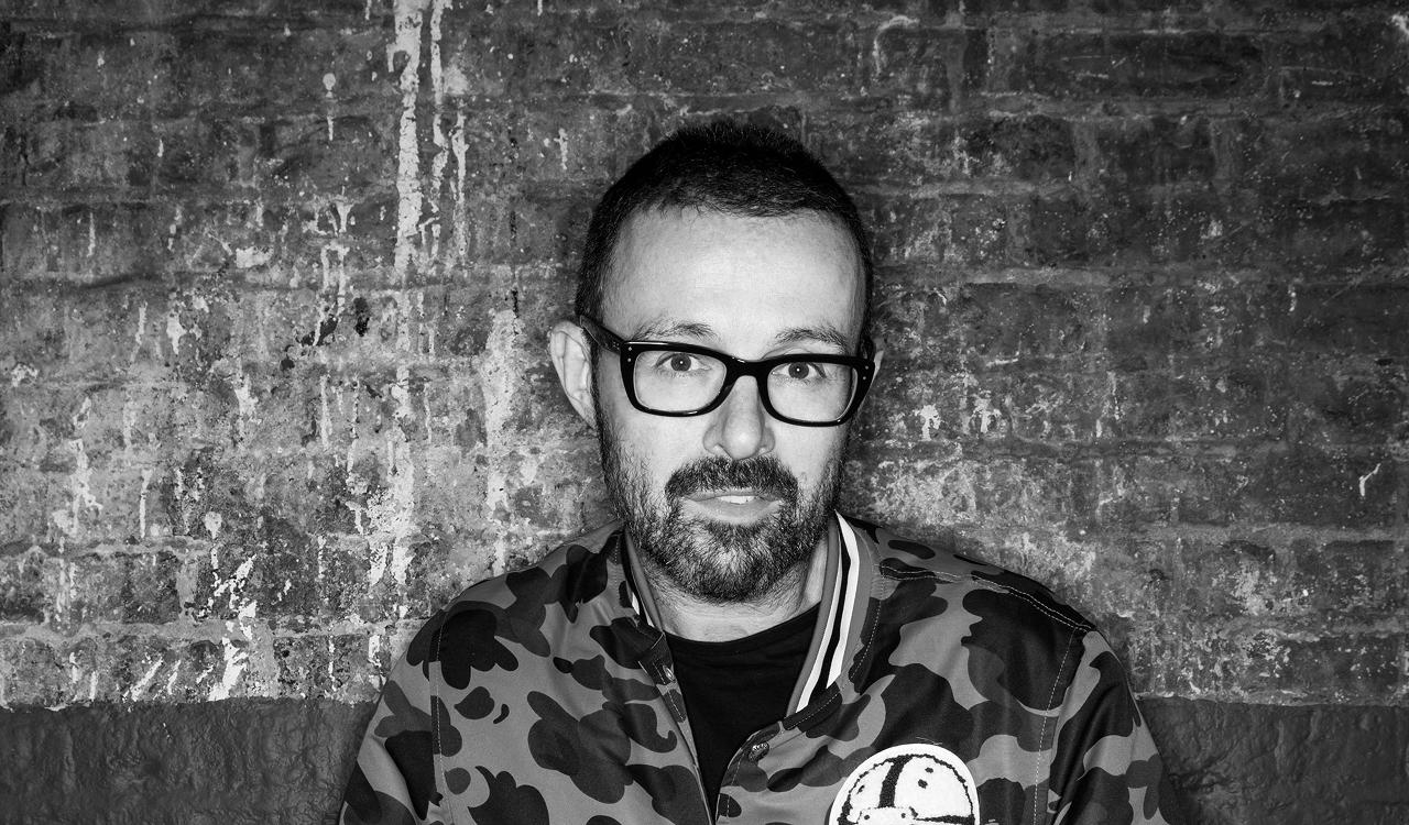 Judge Jules in a black and white portrait against a weathered brick wall, looking at the camera, wearing thick framed glasses and a camouflage bomber jacket.