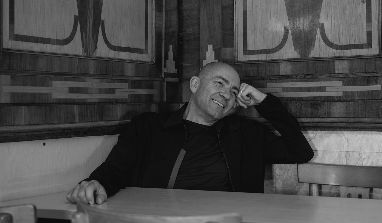 Kenny Thomas portrait. Black and white photo of a smiling bald man in a dark jacket and t-shirt, leaning on one hand while seated at a table in a booth with decorative inlaid wood panelling behind him.