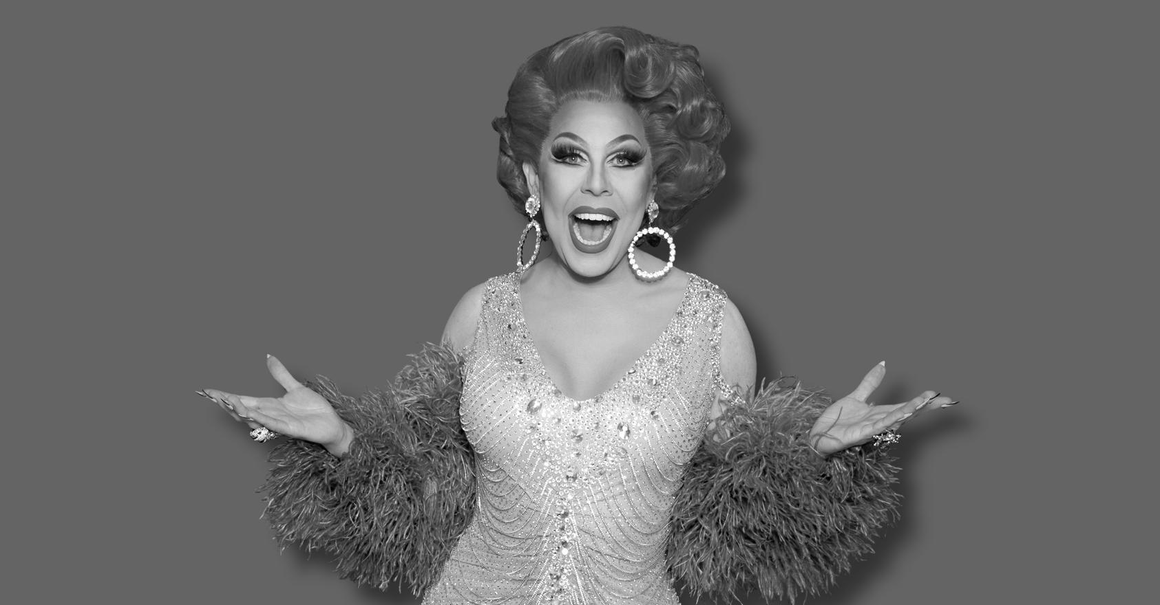 La Voix studio portrait. Black and white photo of the drag performer smiling with mouth open and arms spread wide, wearing a voluminous curled updo, large hoop earrings and a beaded, sequinned gown with feathered cuffs, against a plain grey background.