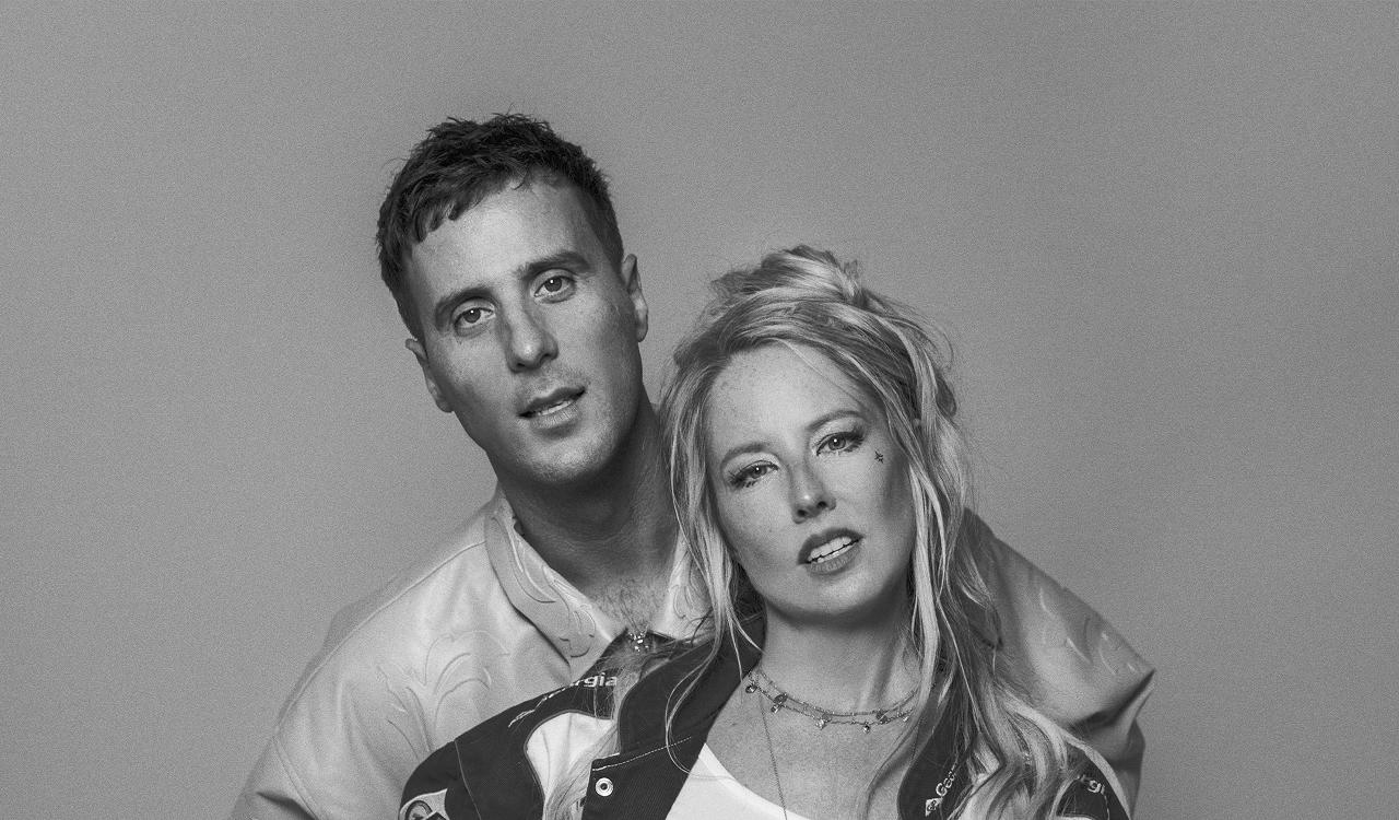 Lovely Laura and Ben Santiago in a black and white portrait against a plain grey background, a man standing behind a woman with long blonde hair, both looking at the camera, wearing jackets and layered necklaces.