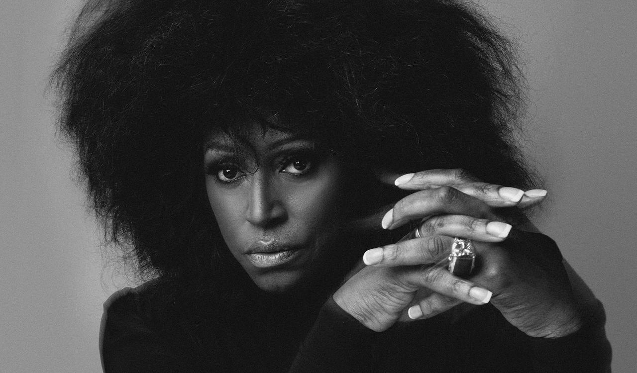 Mica Paris studio portrait. Black and white close-up of a woman with voluminous hair looking directly at the camera, hands clasped beside her face showing long nails and a large statement ring, against a plain grey background.