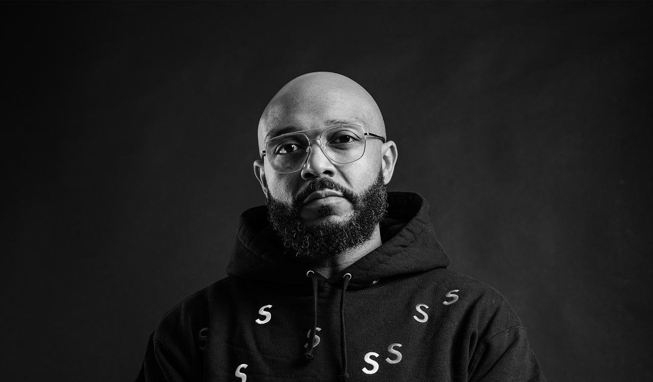Mista Jam in a black and white portrait against a dark background, looking at the camera, wearing aviator glasses and a black hoodie printed with white letter S motifs.