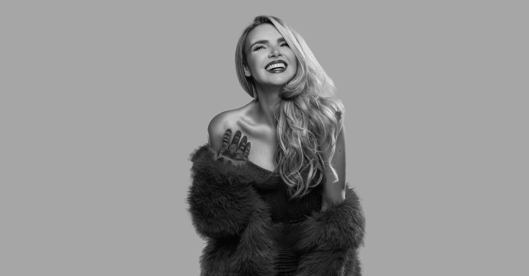 Nadine Coyle studio portrait. Black and white photo of a smiling woman with long, wavy blonde hair, wearing a sparkly strapless dress with a faux fur coat slipping off her shoulders and one gloved hand raised to her chest, against a plain grey background.
