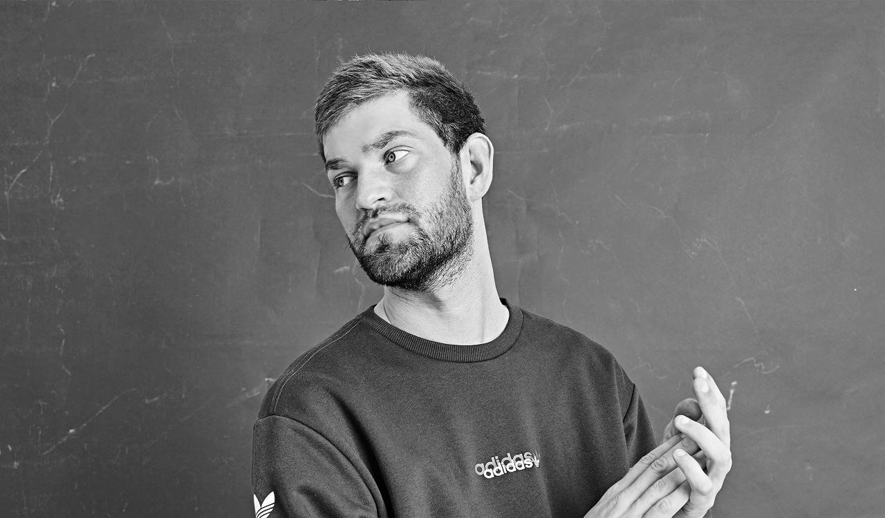 Nathan Dawe in a black and white portrait against a textured grey wall, glancing sideways with hands clasped, wearing a dark adidas sweatshirt.