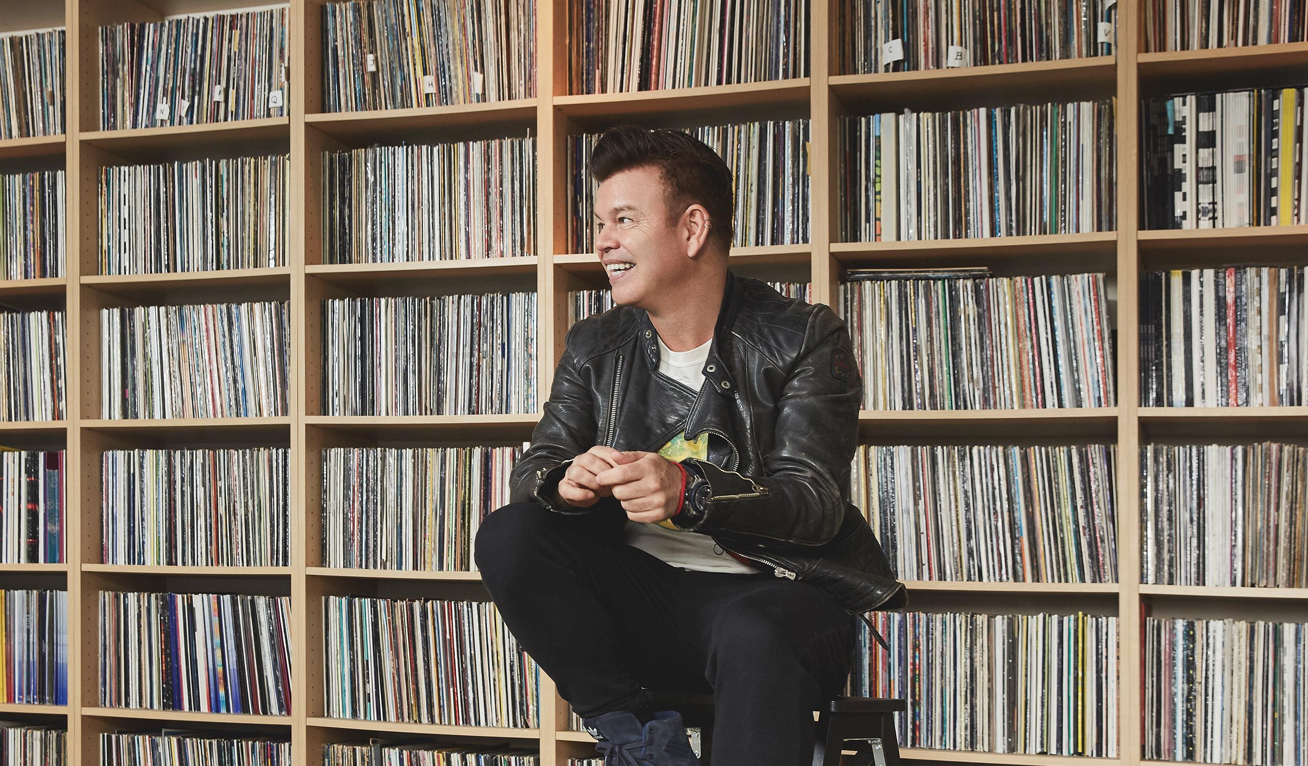 Paul Oakenfold sat in front of shelves full of vinyl records, smiling.