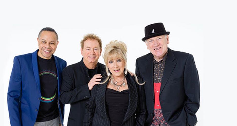 four band members smiling against a white background. From left to right — a man in a blue blazer, a man in a black jacket, a woman in a black outfit with a silver necklace, and a man in a black hat and red-patterned shirt.
