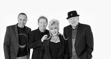 Shakatak group portrait. Black and white photo of the four band members standing together and smiling: three men in dark jackets, one wearing a trilby, and a woman with short blonde hair and a necklace, against a plain white background.