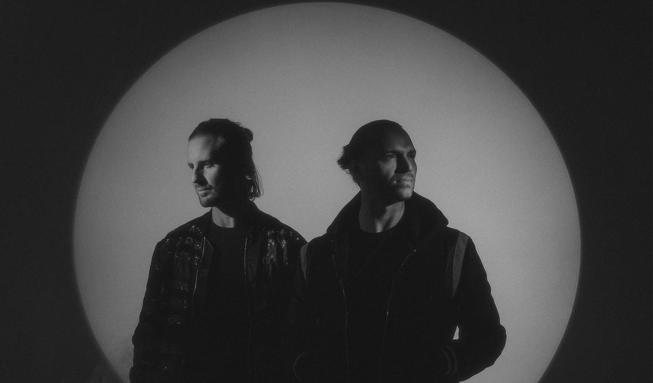 Sigma in a moody black and white photo, two men in dark jackets standing side by side, silhouetted against a large circular spotlight.
