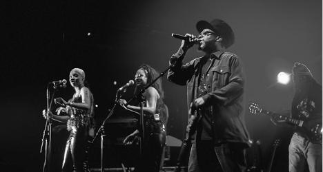 Soul II Soul performing live on stage. Black and white photo of a lead vocalist in a dark cap, glasses and jacket singing into a microphone, with two backing singers at microphone stands to the left and a guitarist on the right, against a dark background with a single spotlight.