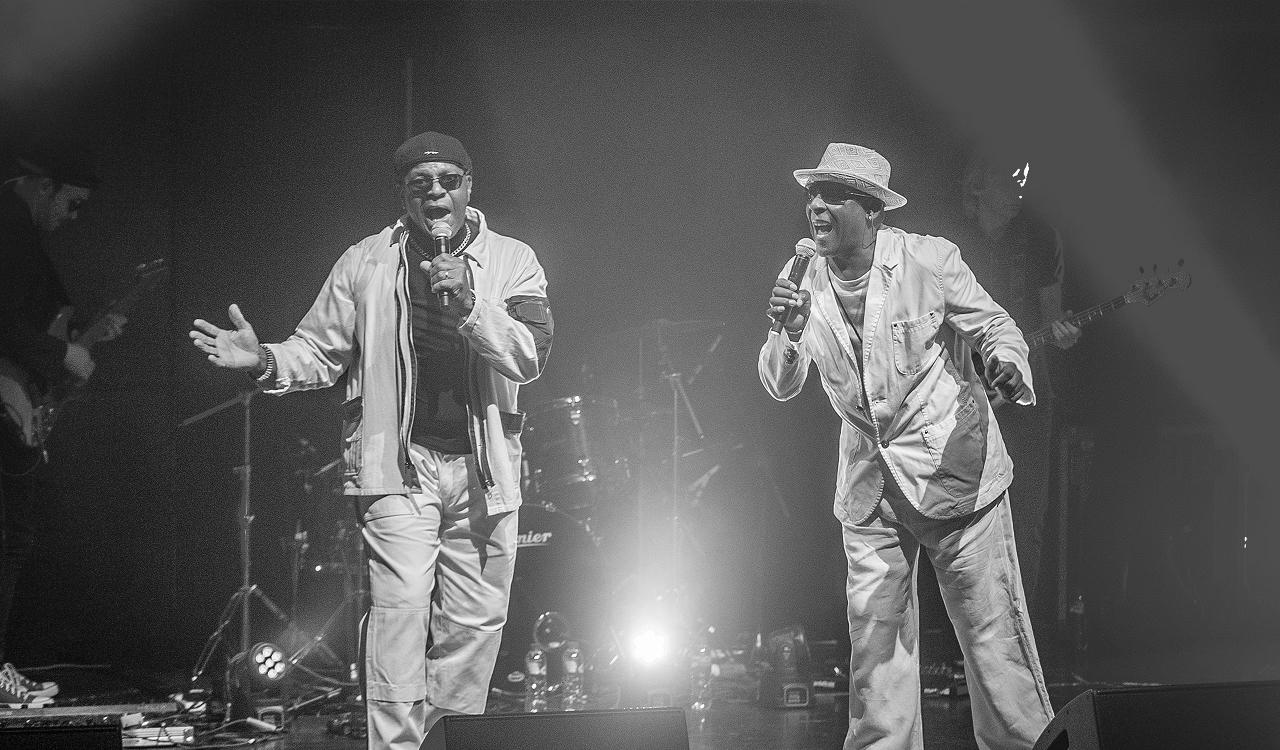 The Real Thing performing live on stage. Black and white photo of two vocalists in light-coloured jackets and trousers singing into handheld microphones, one in a dark beanie with an arm outstretched, the other in a patterned trilby leaning forward. Guitarists and a drum kit sit in the dark background, with a bright stage light shining between the singers.