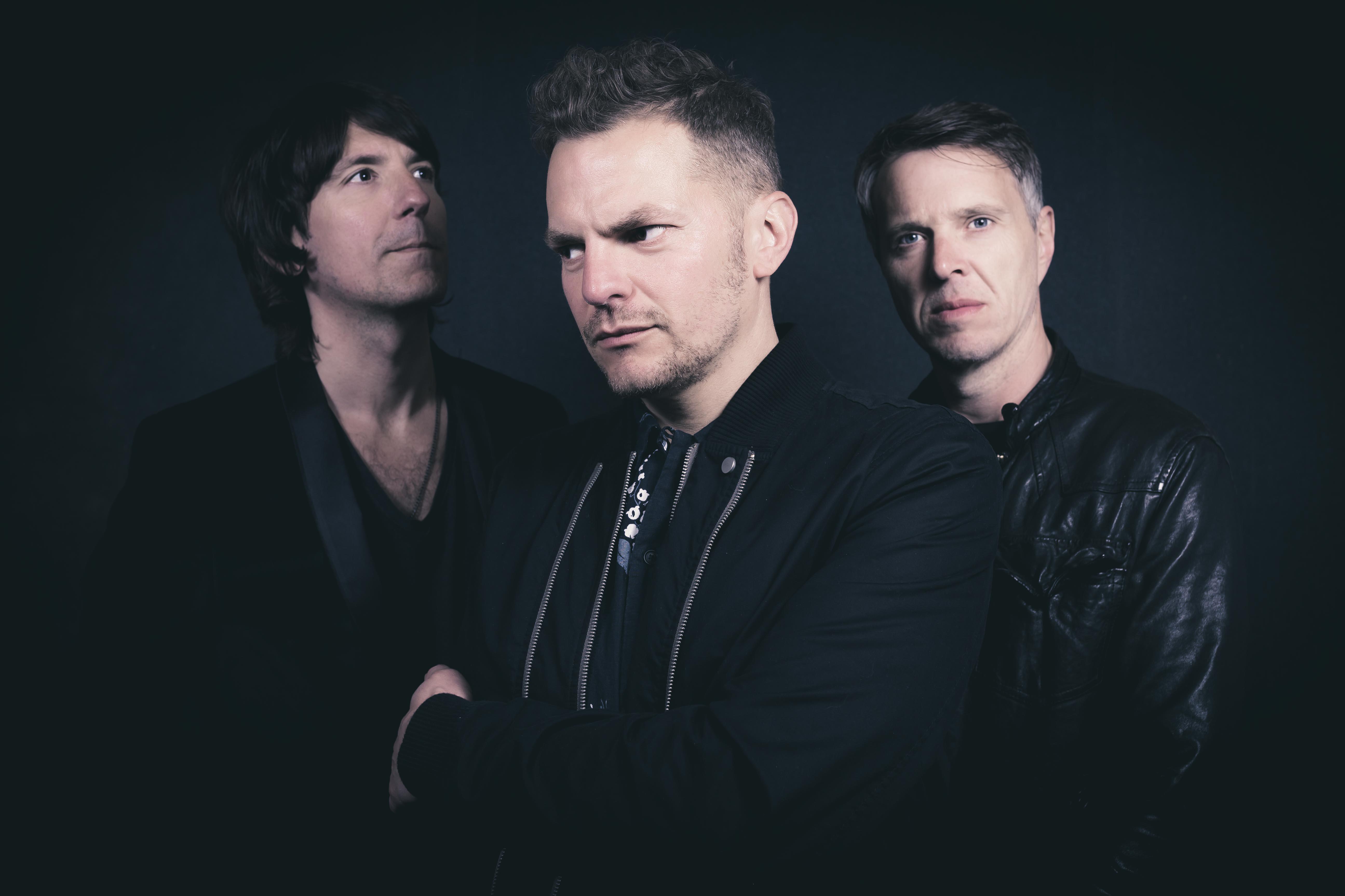 three members of Toploader posed against a dark background, all dressed in black jackets, with the central figure facing forward and the other two positioned slightly behind on either side