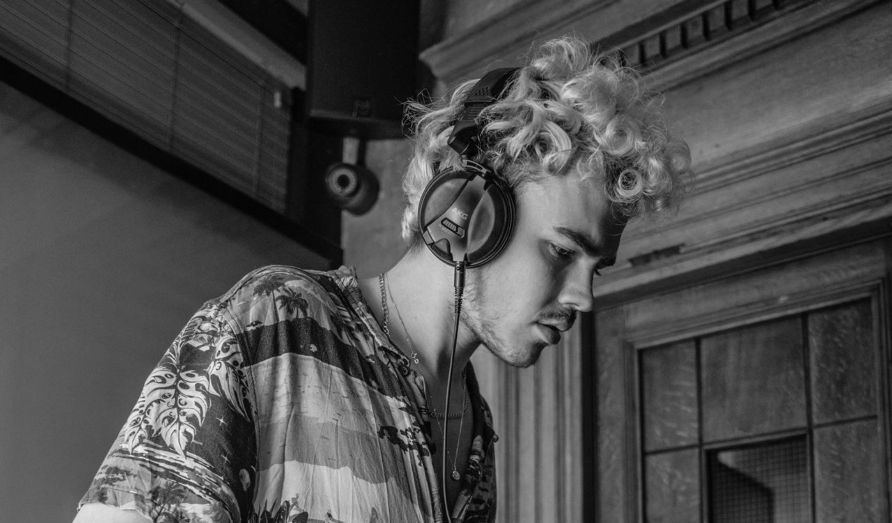 Woody Cook DJing in black and white, wearing AKG headphones and a tropical leaf print shirt, looking down at the decks with curly hair, inside a venue with wood panelled walls and a speaker overhead.
