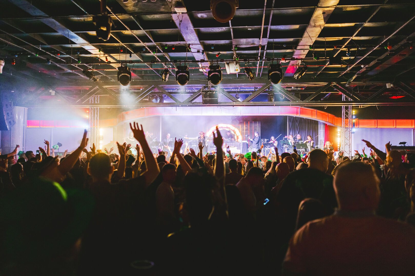A crowd dancing with their hands in the air, in the background is stage with smoke and lights.