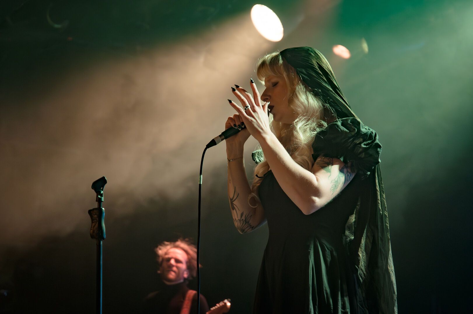 A singer in a dark dress performs into a handheld microphone under green stage lighting, one hand raised expressively, with a guitarist blurred in the background.