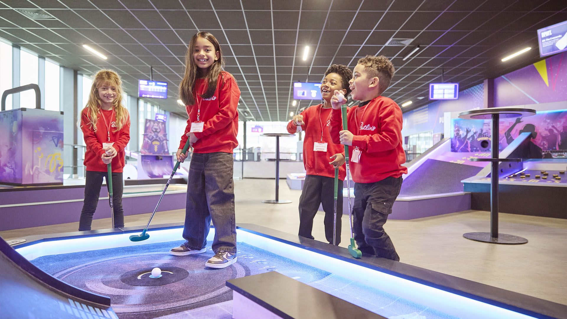 Four children in red sweatshirts stand around an indoor mini-golf hole, laughing and holding putters as a ball rests near the cup.