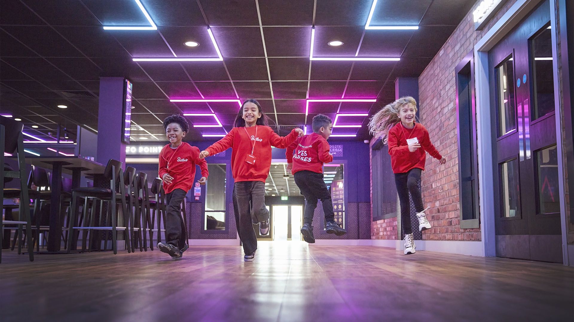 Four children in red sweatshirts run and laugh through an indoor entertainment area with neon ceiling lights.