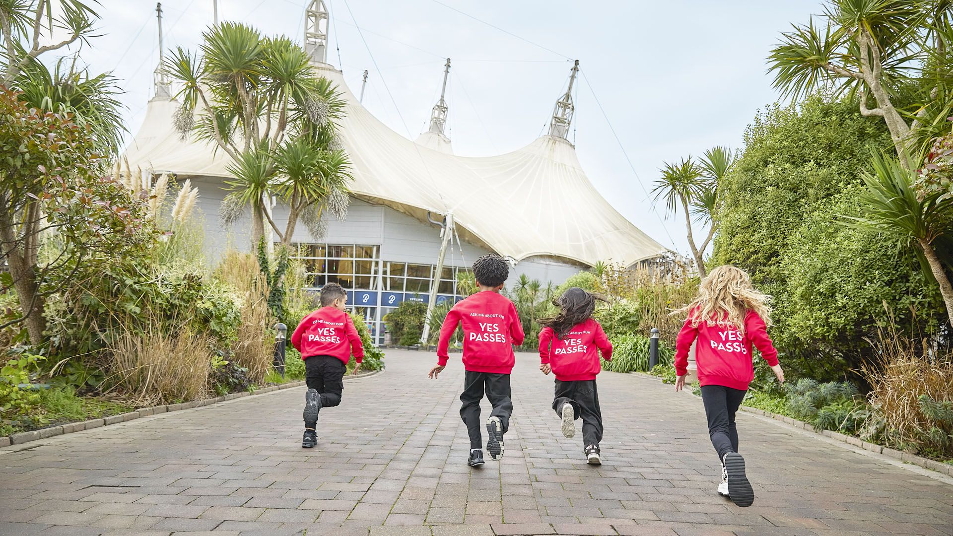 Four children in red “YES PASSES” sweatshirts run along a path toward a large white canopy building surrounded by greenery.