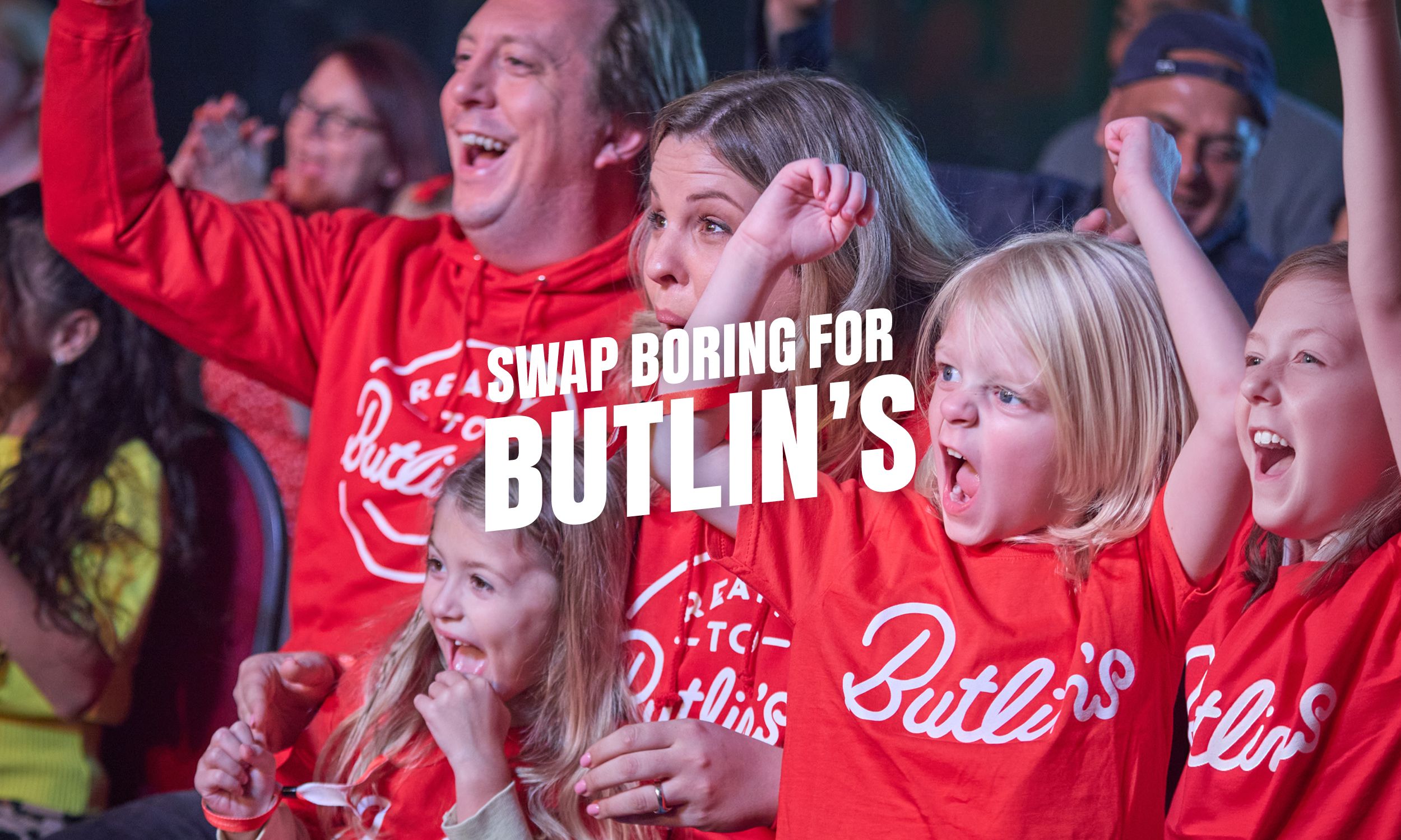 A family wearing matching red Butlin’s T-shirts cheers and smiles in a lively show audience, with children raising their arms in excitement. The text “Swap boring for Butlin’s” appears over the image.