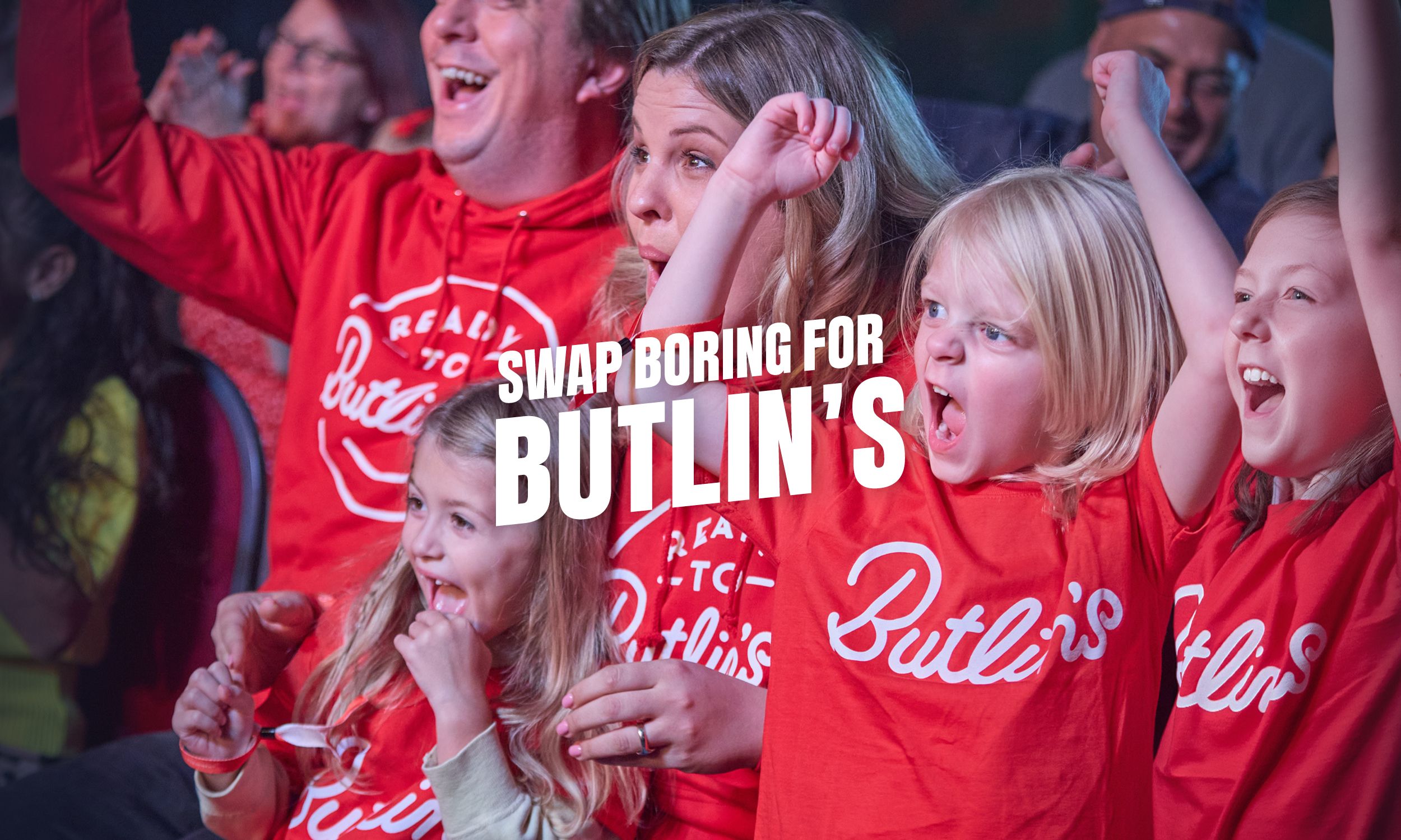 A family wearing matching red Butlin’s T-shirts cheers and smiles in a lively show audience, with children raising their arms in excitement. The text “Swap boring for Butlin’s” appears over the image.