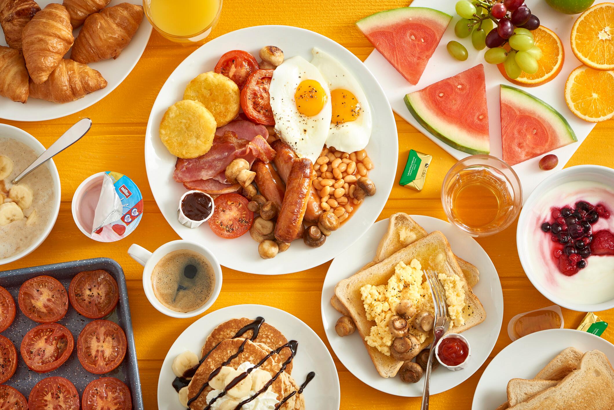 A table filled with breakfast meals consisting of pancakes, breads, fruits, sausages, eggs, bacons and eggs.