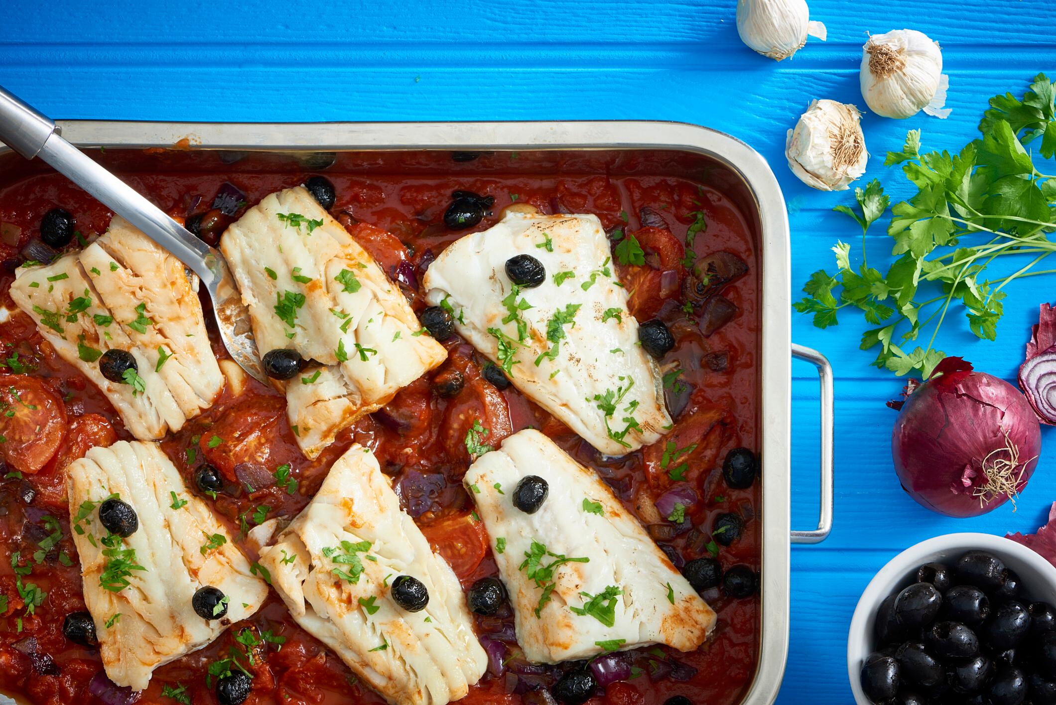 A table with a tray of food filled with fish fillet soaked in tomato sauce and olives.
