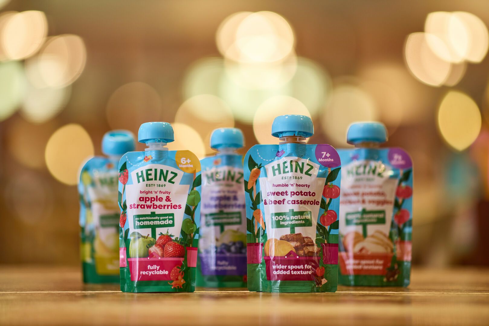 A focused image of Heinz baby food pouches on a table, with two flavours prominently shown in the foreground: “Apple & Strawberries” and “Sweet Potato & Beef Casserole.” The background is softly blurred with warm lights.