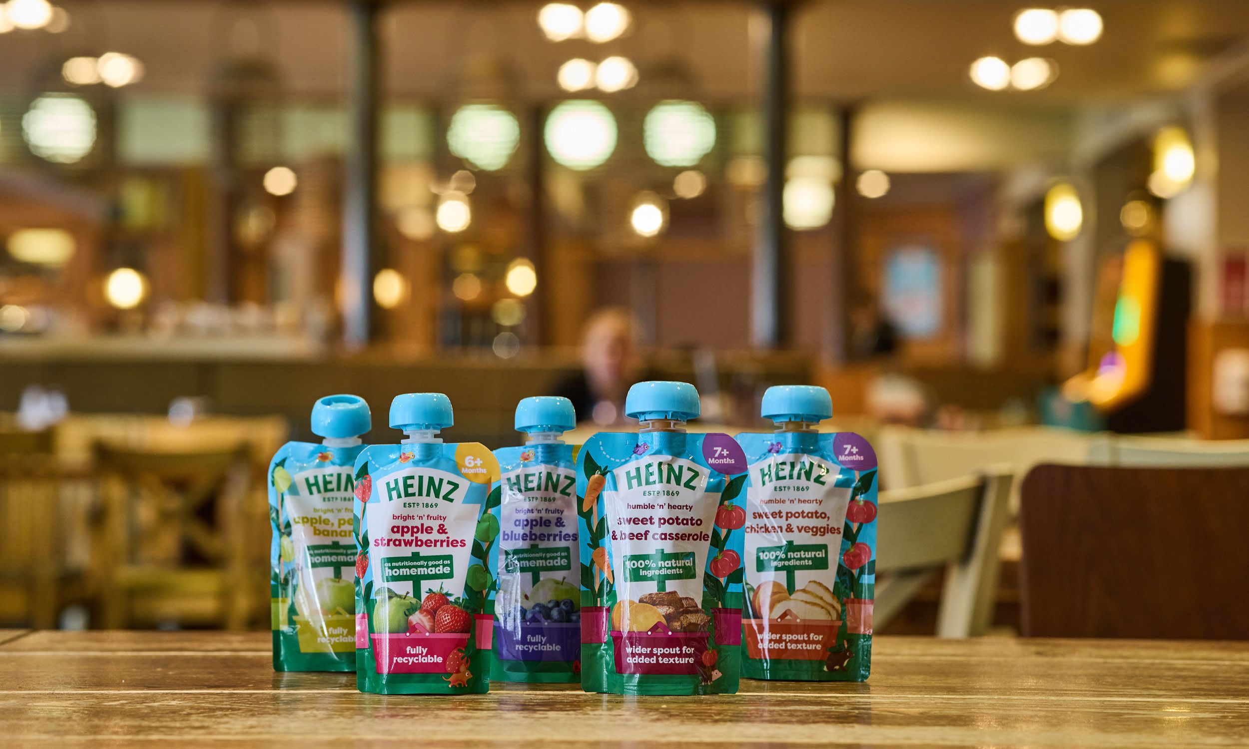 A wide shot of six Heinz baby food pouches neatly lined up on a wooden surface, featuring flavours like “Apple, Pear & Banana,” “Apple & Blueberries,” and “Sweet Potato, Parsnip & Veggies.” Blurred restaurant-style lighting glows in the background.