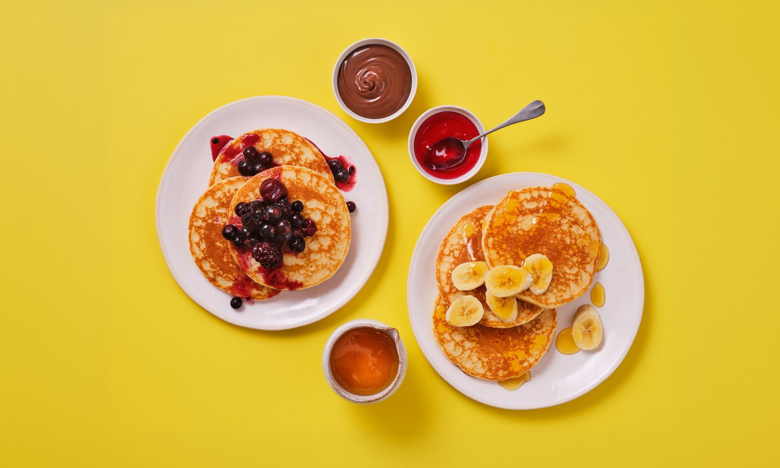 A flatlay of two plates of pancakes on a bright yellow background. One stack is topped with mixed berry compote and blueberries, the other with sliced banana and maple syrup. Accompaniments include a small jug of maple syrup, a pot of chocolate spread, and a pot of strawberry jam with a spoon.