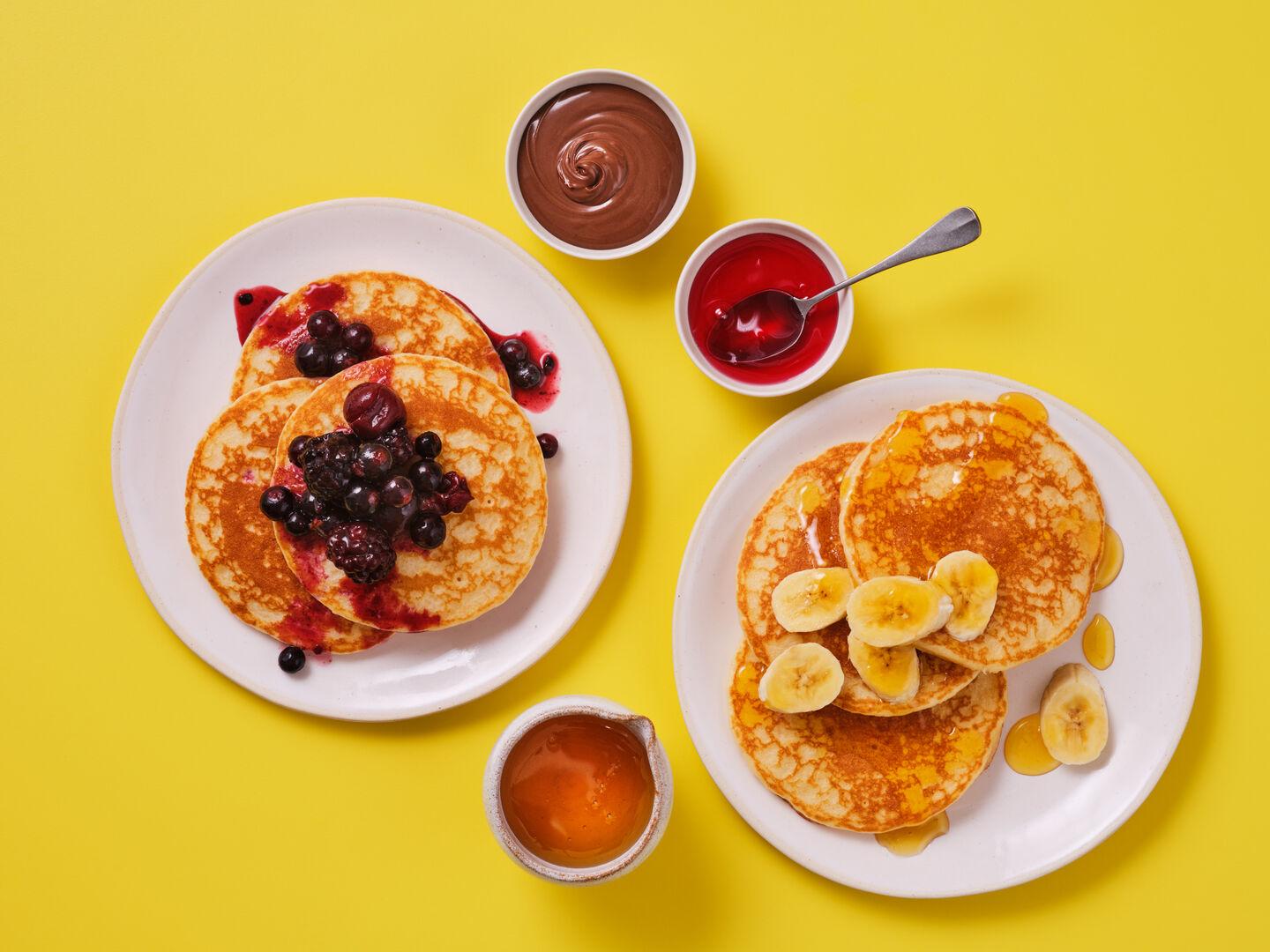Two plates of pancakes on a yellow background, one topped with mixed berries, the other with sliced banana and syrup, alongside pots of chocolate sauce, fruit compote and honey.