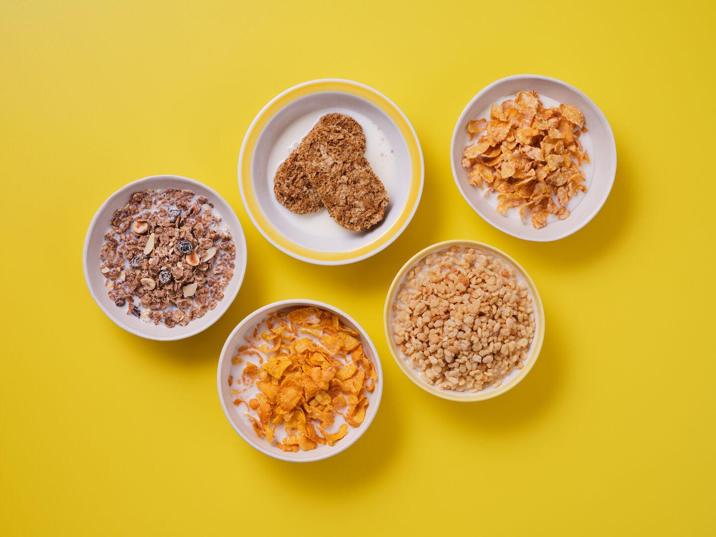 Five bowls of cereal with milk on a yellow background, including muesli, wheat biscuits, cornflakes, frosted flakes and rice cereal.