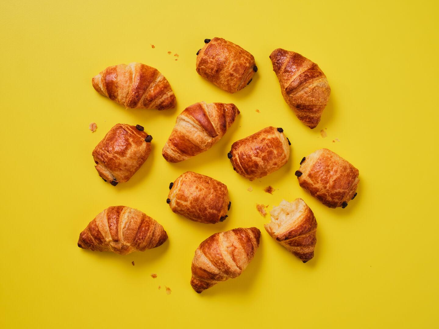 A selection of croissants and pain au chocolat on a yellow background, with one croissant broken open.