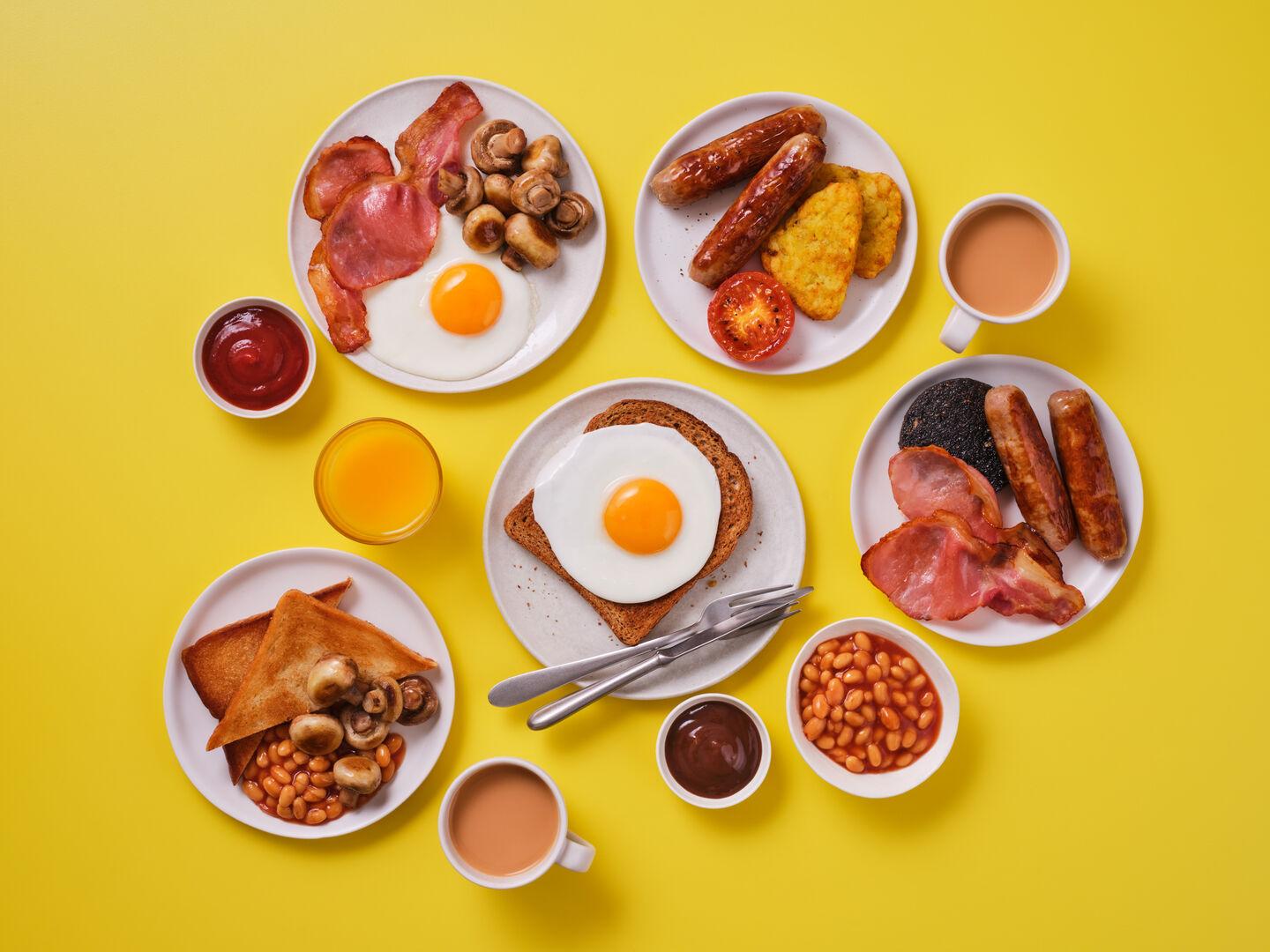 A full English breakfast spread on a yellow background, including fried egg, bacon, mushrooms, sausages, hash browns, grilled tomato, black pudding, beans, toast, ketchup, tea and orange juice.