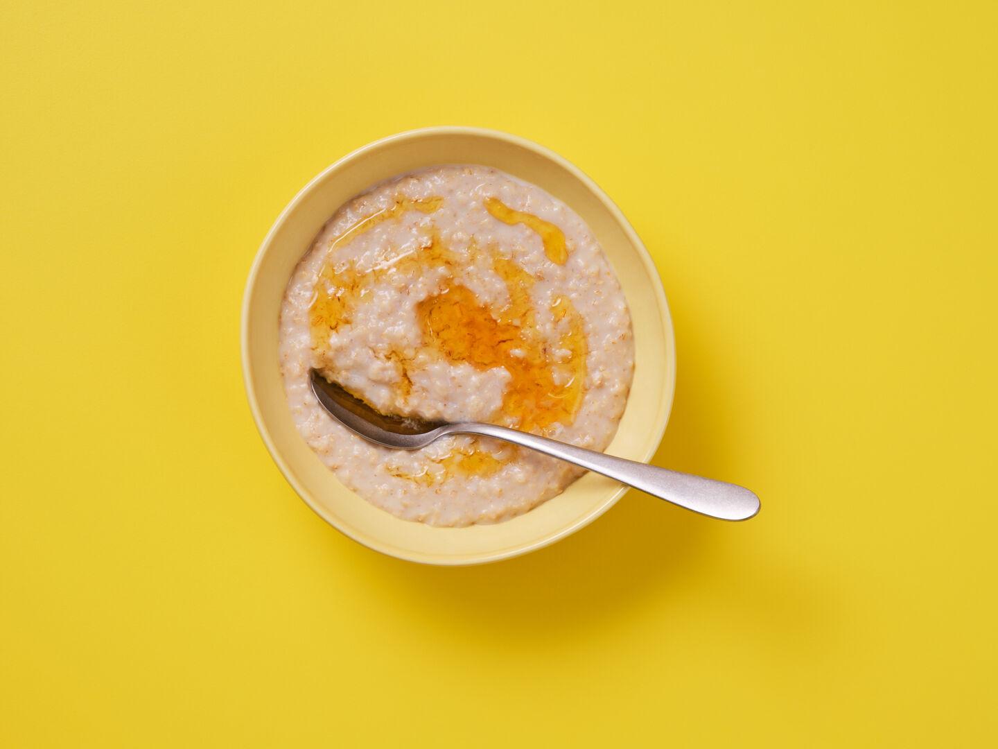 A bowl of porridge drizzled with honey, with a spoon, on a yellow background.