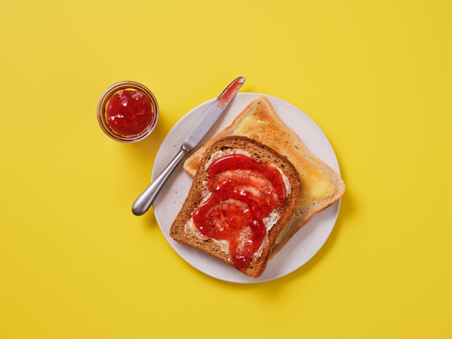 Two slices of buttered toast on a plate, one topped with strawberry jam, with a knife and a small pot of jam alongside, on a yellow background.