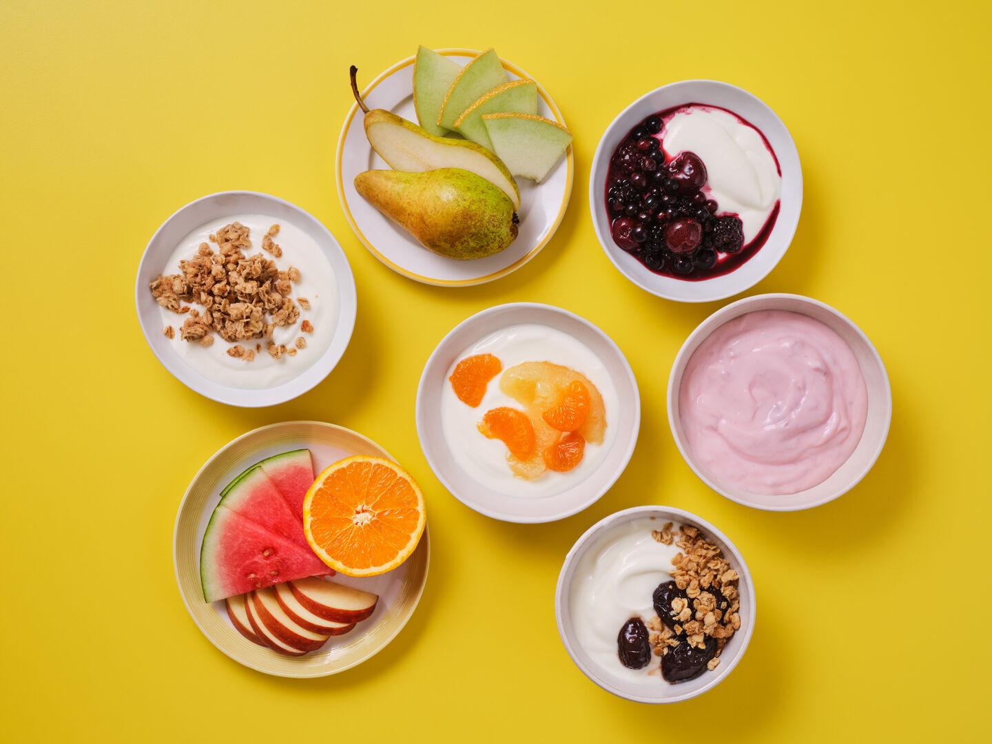 A selection of fresh fruit and yoghurt bowls on a yellow background, including sliced pear and melon, mixed berry compote, yoghurt with granola, mandarin segments, strawberry yoghurt, watermelon and orange slices, and yoghurt with fruit compote and granola.