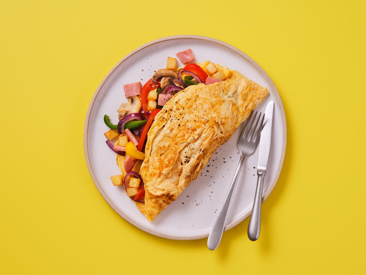 An omelette filled with ham, peppers, onion, mushroom and potato, served with a knife and fork, on a yellow background.