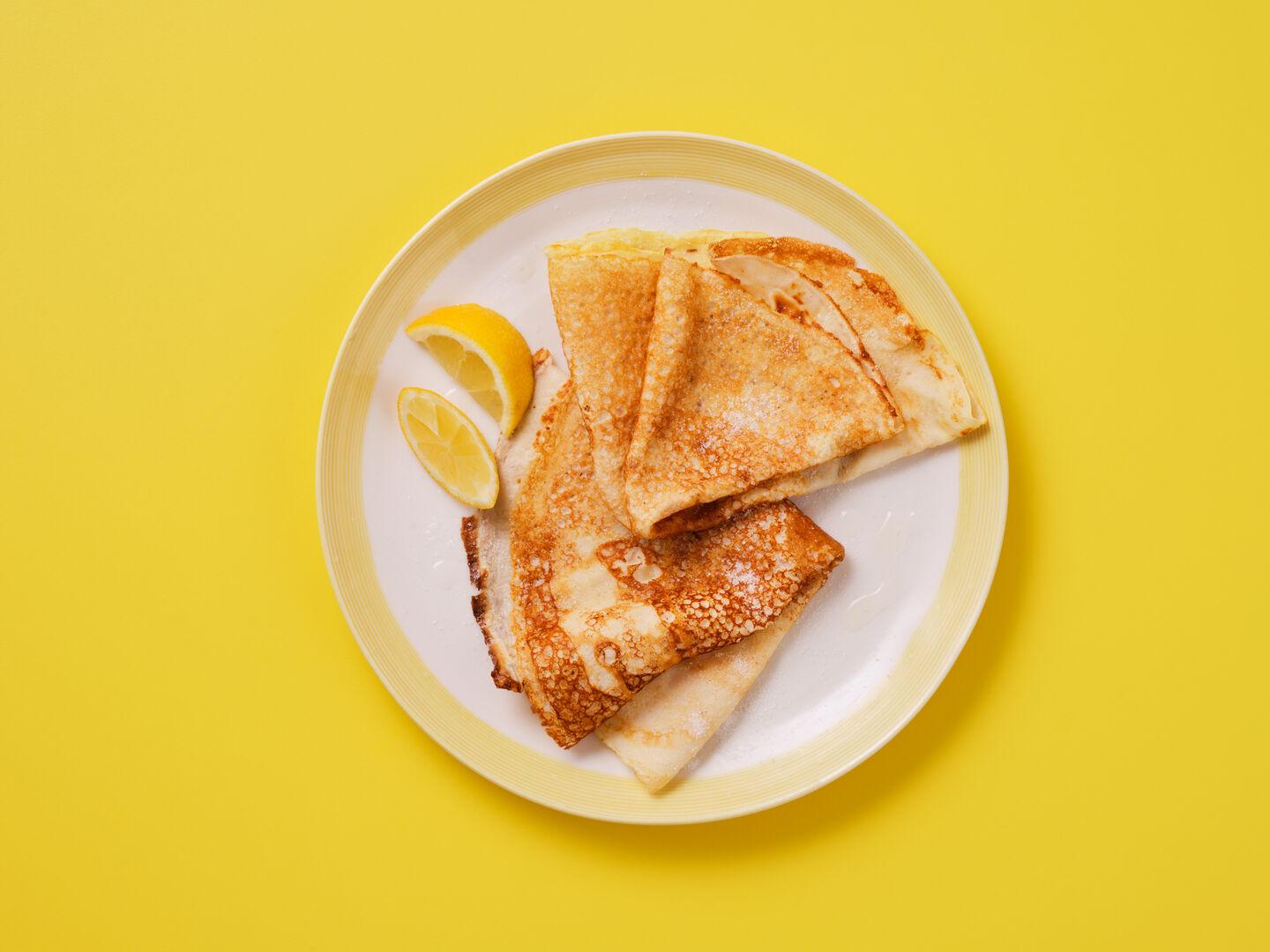 Folded pancakes dusted with sugar, served with lemon wedges on a plate, on a yellow background.