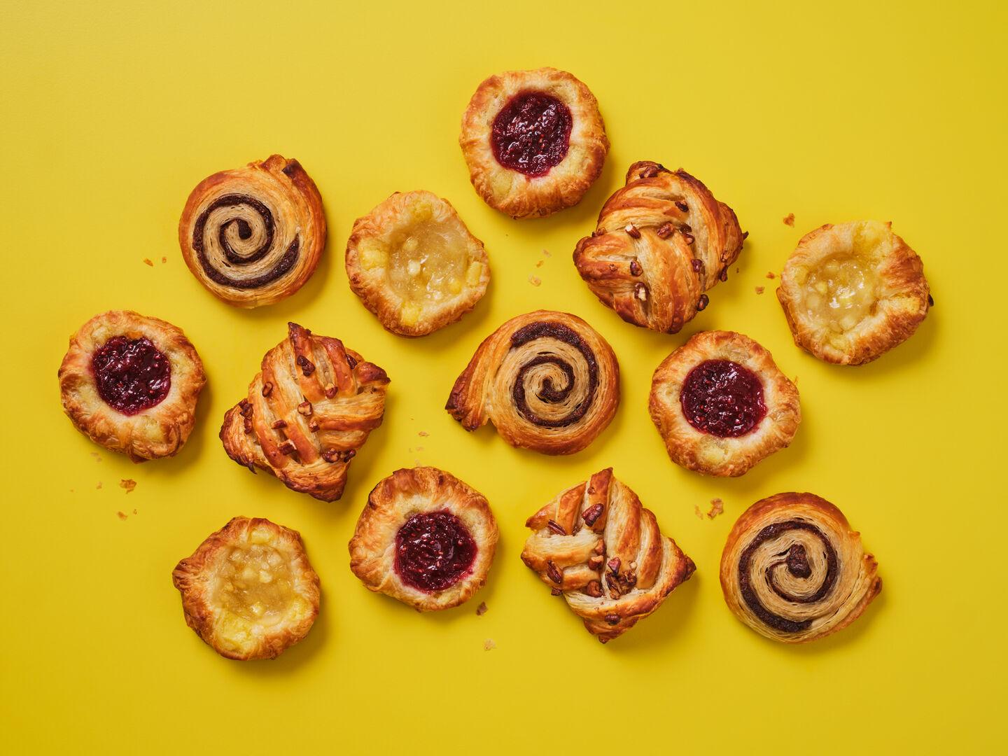 A selection of breakfast pastries on a yellow background, including pain au raisin, pecan plaits, jam-filled and apple Danish pastries.